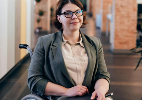 A woman seated in a wheelchair holds a laptop and smiles at the camera